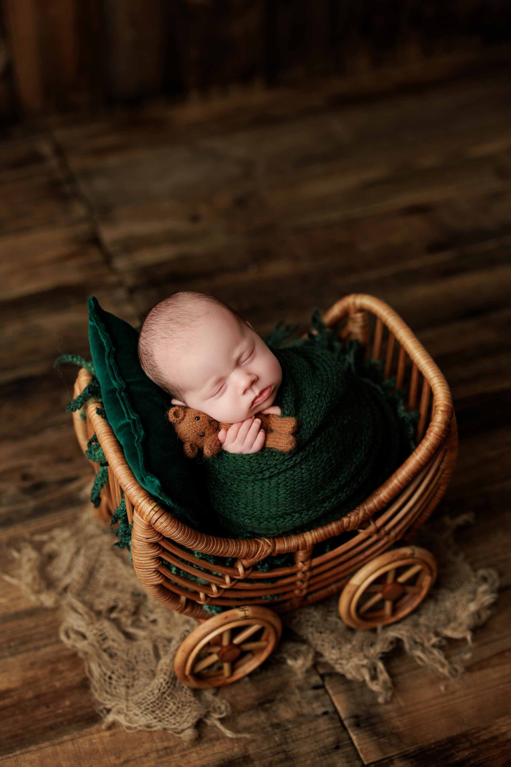 A newborn baby wrapped in a dark green blanket sleeps in a small wicker pram, holding a tiny teddy bear—an adorable moment captured for Adelaide baby photos on a rustic wooden floor with hessian cloth underneath.