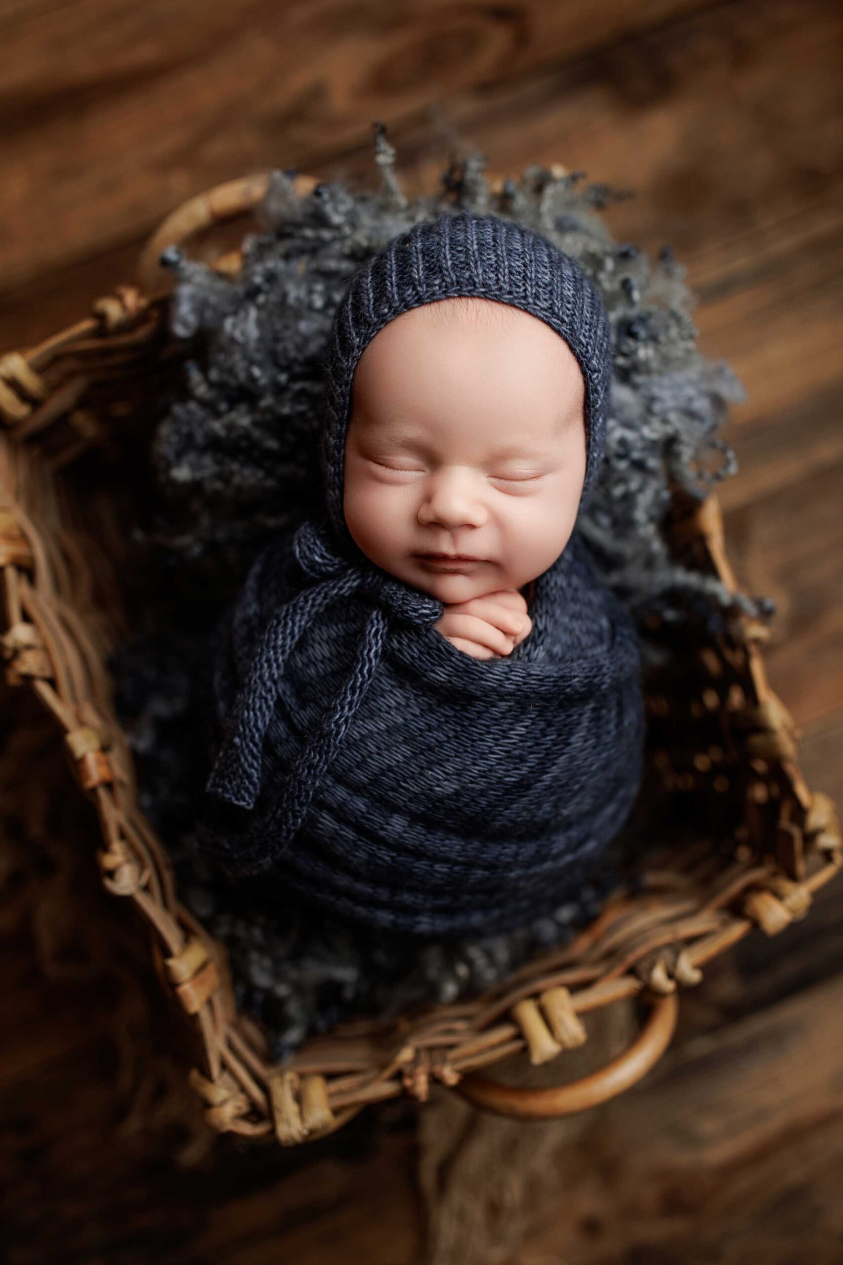 A newborn baby, wrapped in a dark blue knitted blanket and matching bonnet, sleeps peacefully in a wicker basket lined with soft, fluffy fabric on a wooden floor—capturing the charm of Adelaide baby photos.
