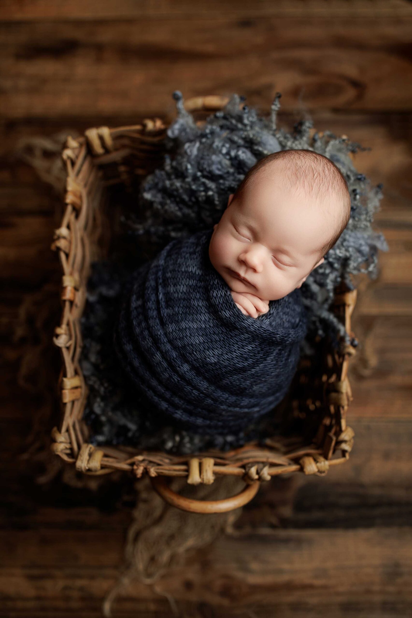 A newborn baby wrapped in a dark blue knitted blanket sleeps peacefully in a wicker basket lined with a textured grey blanket, set on a wooden floor—perfect for timeless Adelaide baby photos.