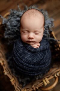 A newborn baby sleeps peacefully, swaddled in a dark blue knitted blanket, lying in a wicker basket lined with soft, textured grey material. The baby’s hands gently rest on their chest—perfect for timeless Adelaide baby photos.