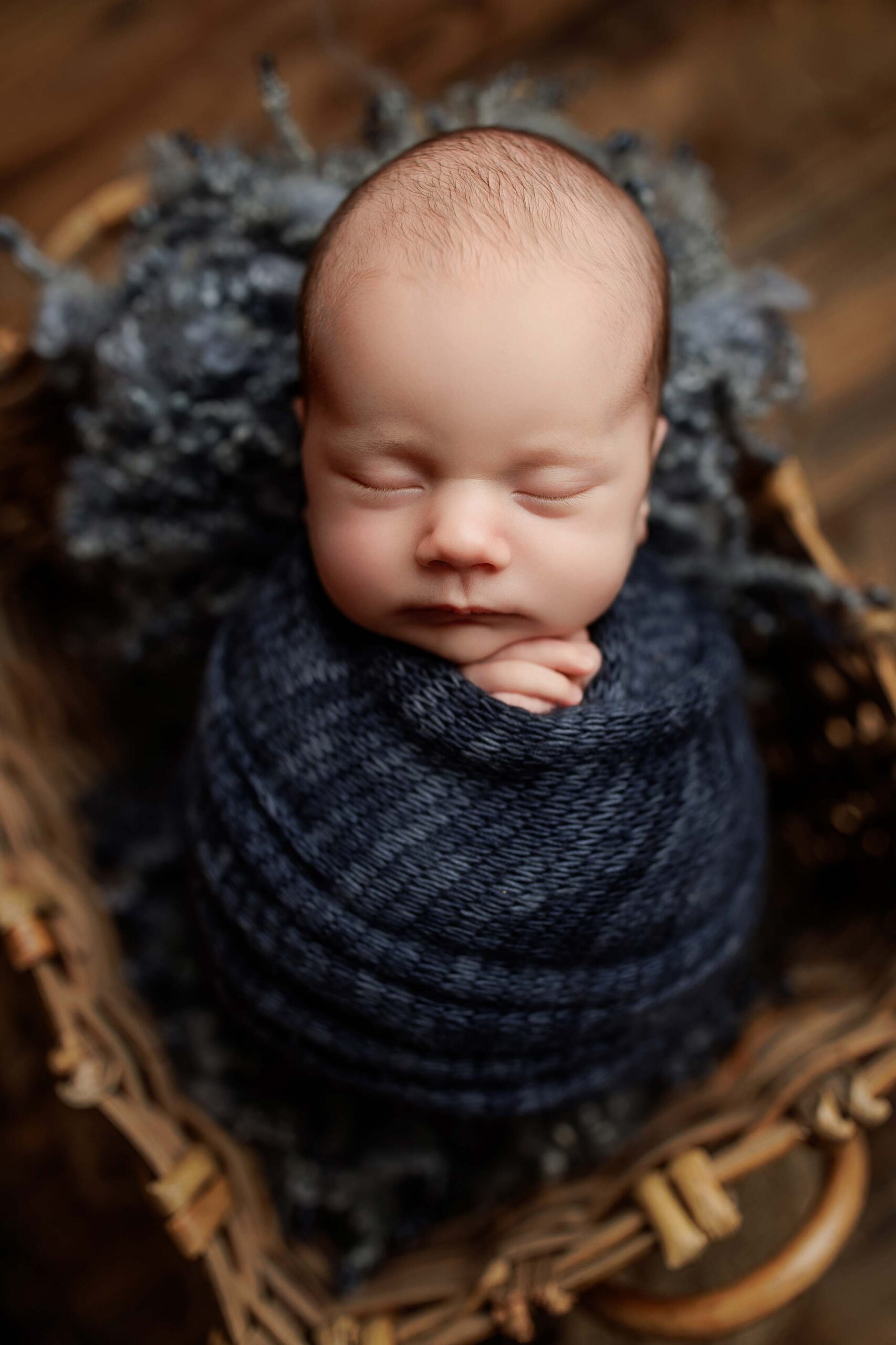 A newborn baby sleeps peacefully, swaddled in a dark blue knitted blanket, lying in a wicker basket lined with soft, textured grey material. The baby’s hands gently rest on their chest—perfect for timeless Adelaide baby photos.