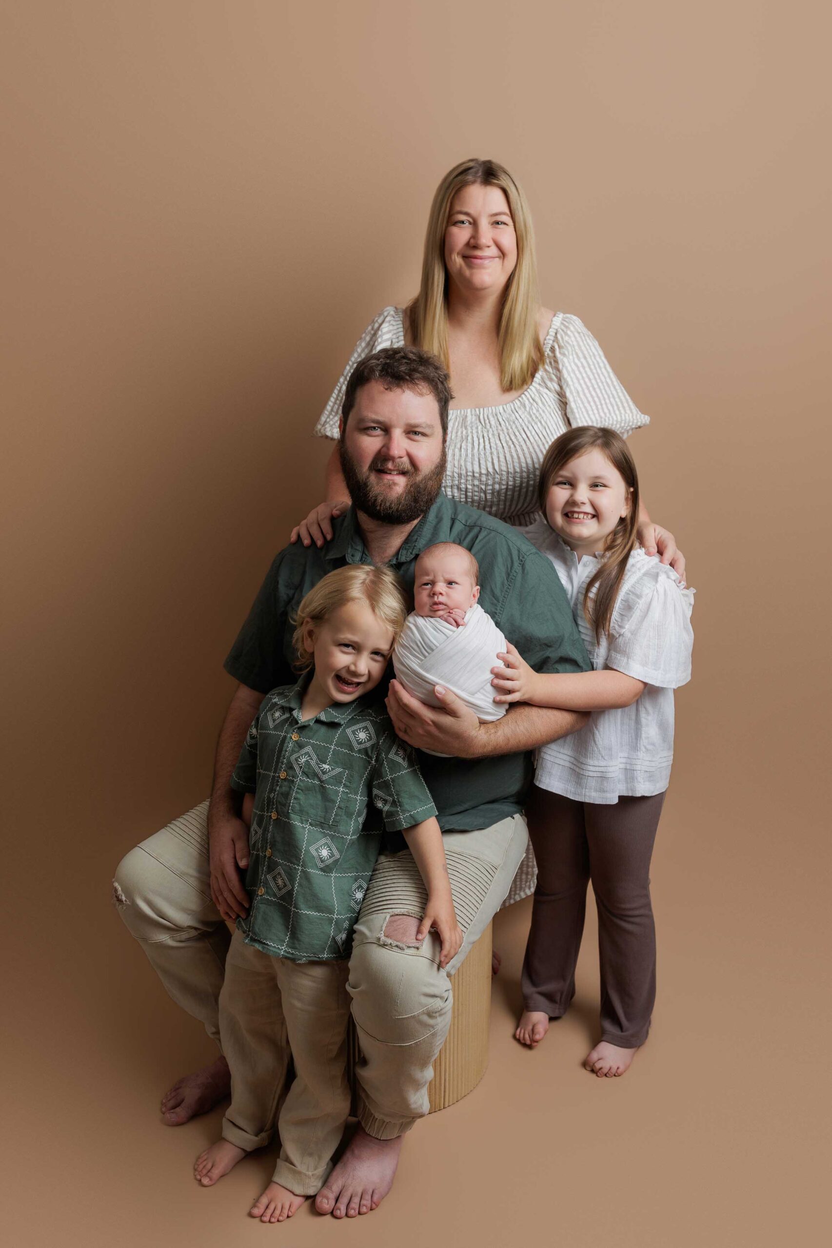 A family of five poses against a beige background for their Adelaide baby photos. The father sits on a stool holding a newborn, with a young boy and girl at his sides, whilst the mother stands behind them, smiling.