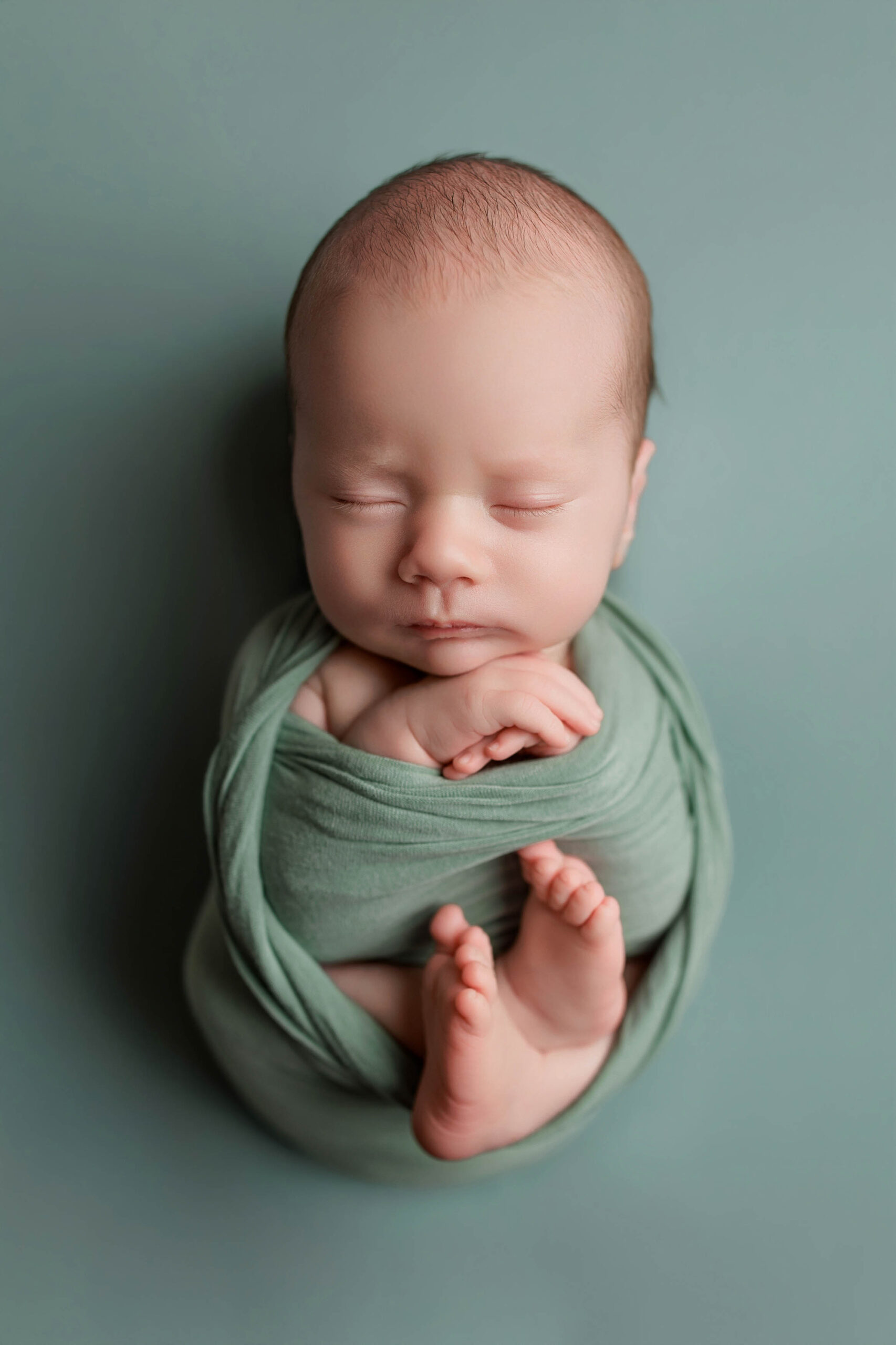 A newborn baby is peacefully sleeping, swaddled in a soft sage green cloth, with their arms gently crossed over their chest and feet tucked up, against a matching light green background—capturing the essence of Adelaide baby photos.