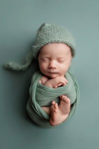 A newborn baby is peacefully sleeping, swaddled in a soft green wrap with a matching knitted hat, lying on a green background. The baby's arms are crossed and tiny feet peek out—perfect for Adelaide baby photos.