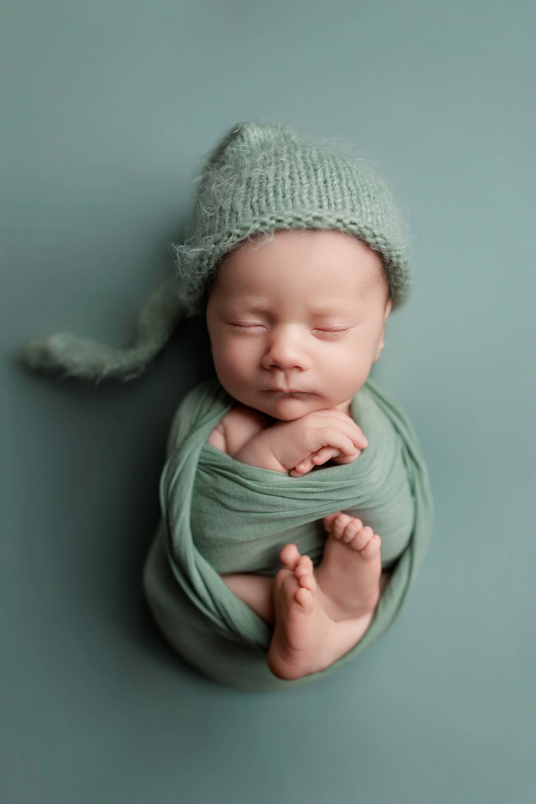 A newborn baby is peacefully sleeping, swaddled in a soft green wrap with a matching knitted hat, lying on a green background. The baby's arms are crossed and tiny feet peek out—perfect for Adelaide baby photos.