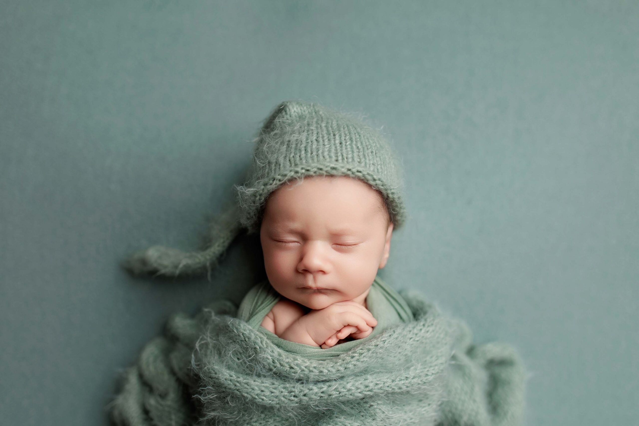 A newborn baby sleeps peacefully, wrapped in a soft mint-green blanket and matching knitted hat with a long tail, set against a pale green background—perfect for capturing sweet Adelaide baby photographs.