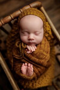 Newborn baby sleeping peacefully in a wooden chair, wrapped in a mustard yellow knitted blanket with matching bonnet—hands folded under chin, tiny feet peeking out—captured from above. Perfect for Adelaide baby photos.