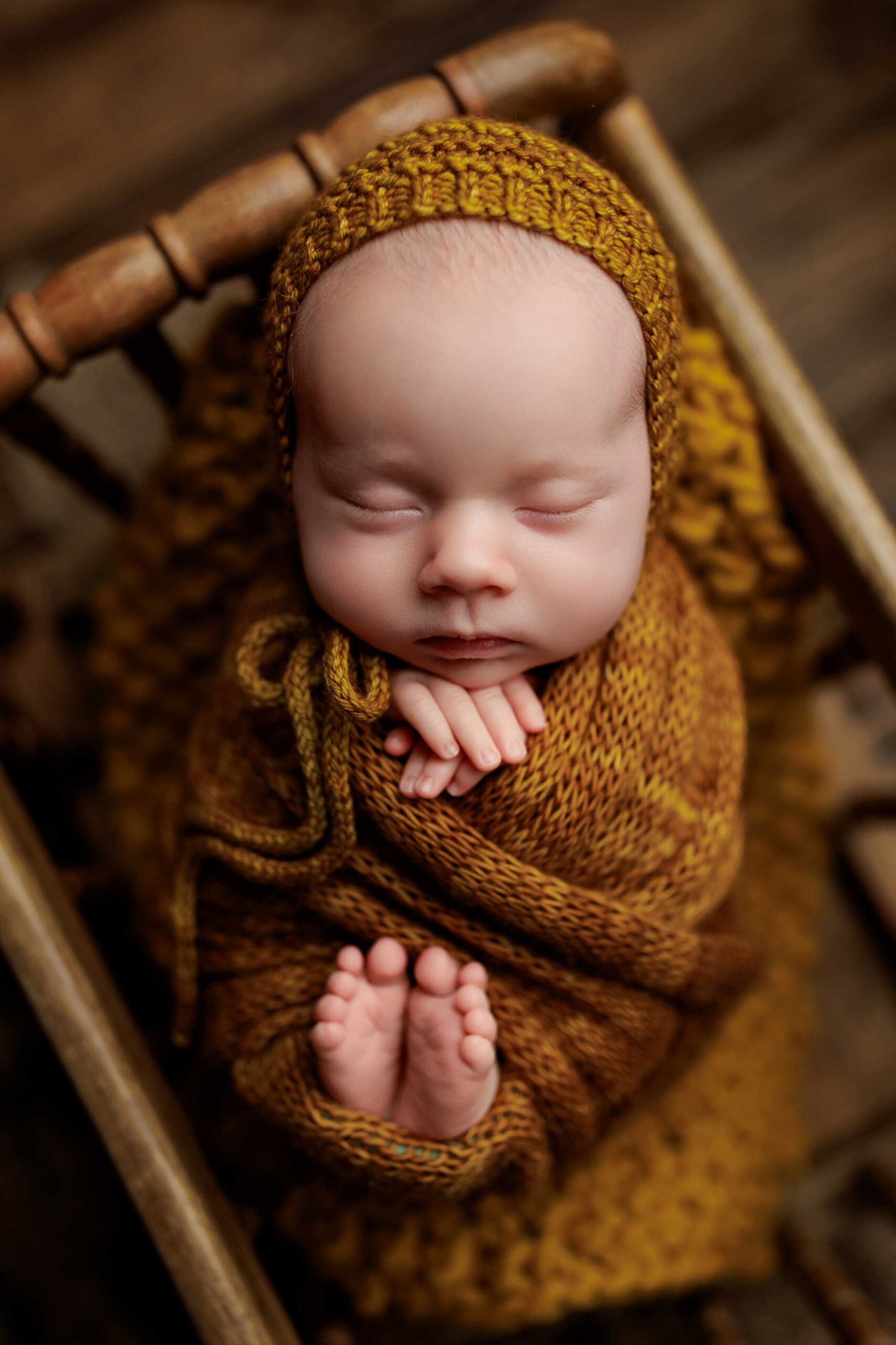 Newborn baby sleeping peacefully in a wooden chair, wrapped in a mustard yellow knitted blanket with matching bonnet—hands folded under chin, tiny feet peeking out—captured from above. Perfect for Adelaide baby photos.