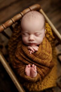 A newborn baby sleeps peacefully in a wooden cot, wrapped in a cosy brown knitted blanket with their hands and feet gently crossed—an adorable moment captured in classic Adelaide baby photos against a softly blurred, warm-toned background.