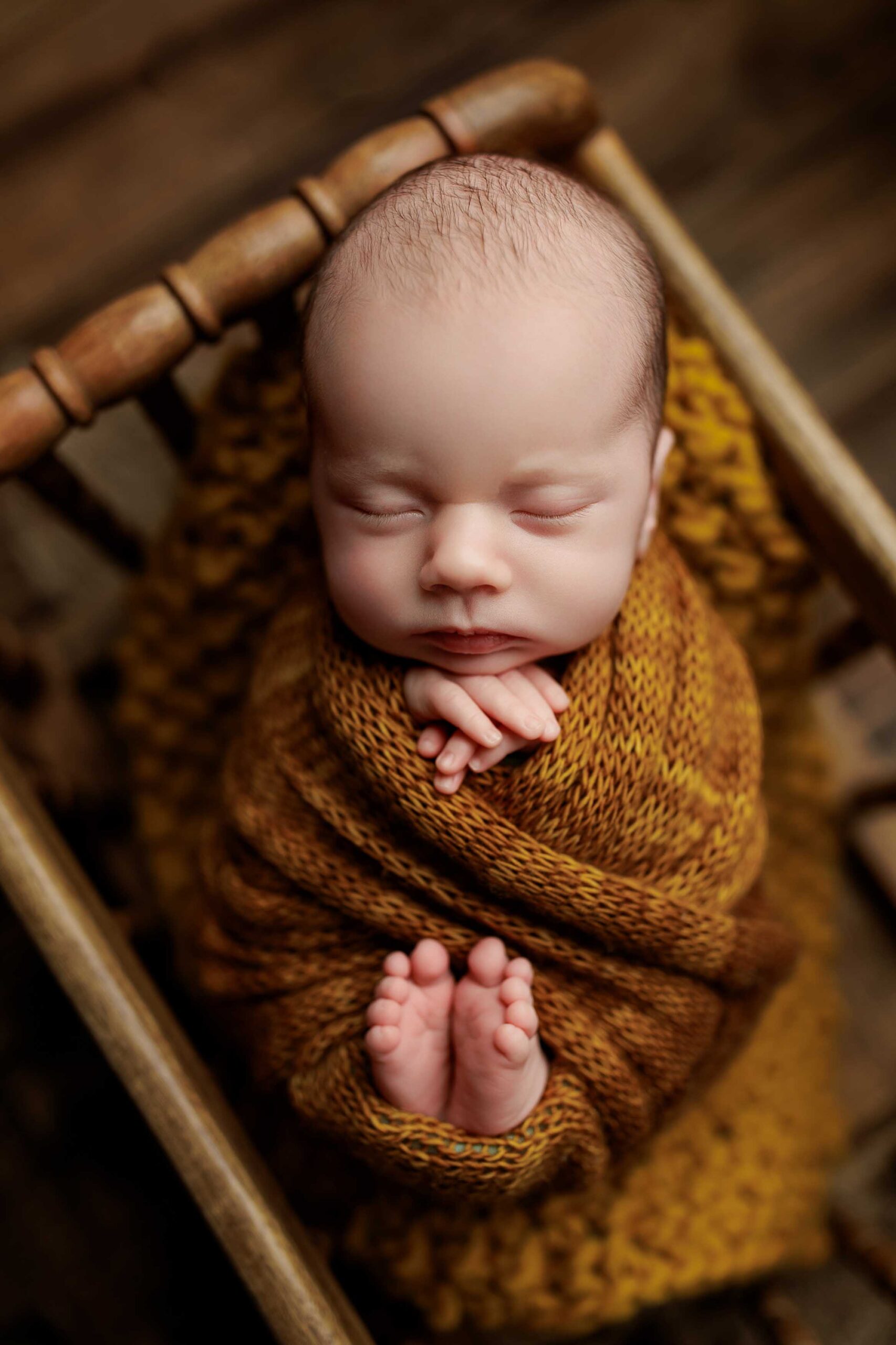 A newborn baby sleeps peacefully in a wooden cot, wrapped in a cosy brown knitted blanket with their hands and feet gently crossed—an adorable moment captured in classic Adelaide baby photos against a softly blurred, warm-toned background.