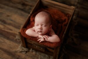 A newborn baby sleeps peacefully with arms folded under its chin, resting on a soft brown blanket inside a rustic wooden box on a wooden floor—capturing the charm of Adelaide baby photos.