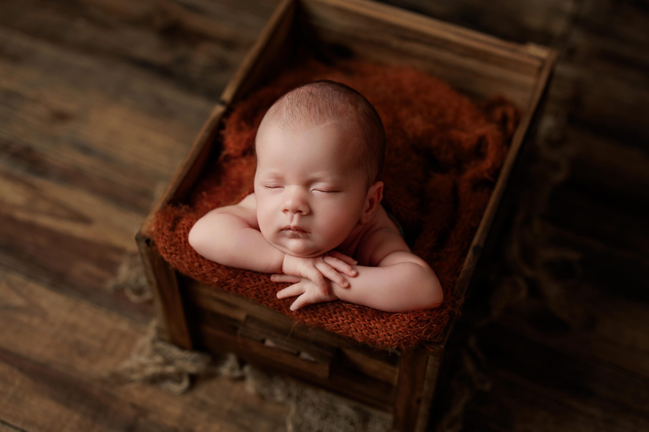 A newborn baby sleeps peacefully with arms folded under its chin, resting on a soft brown blanket inside a rustic wooden box on a wooden floor—capturing the charm of Adelaide baby photos.