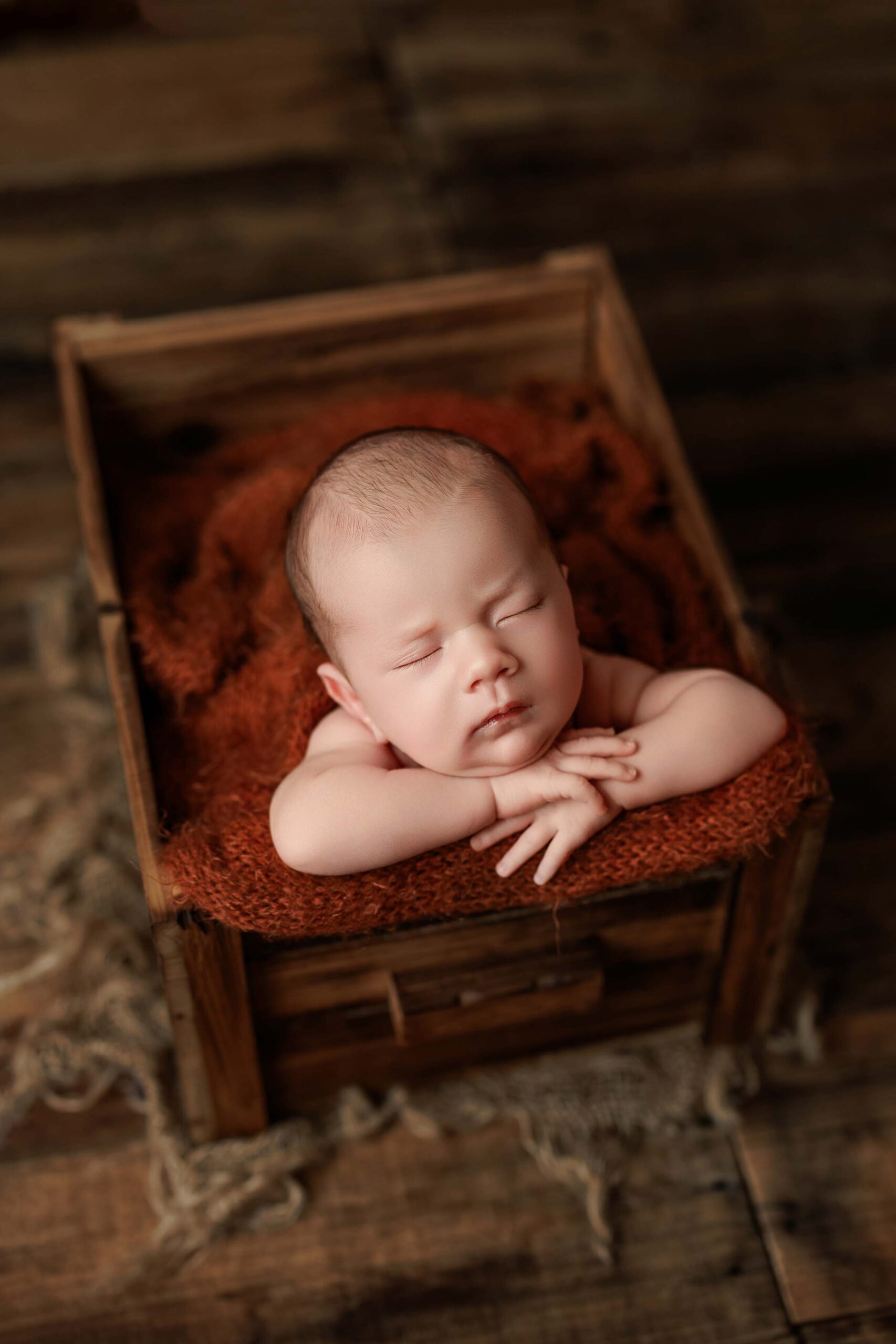 A baby sleeps peacefully with arms folded under its chin, resting on a soft brown blanket inside a rustic wooden box on a wooden floor—capturing the charm of Adelaide baby photos.