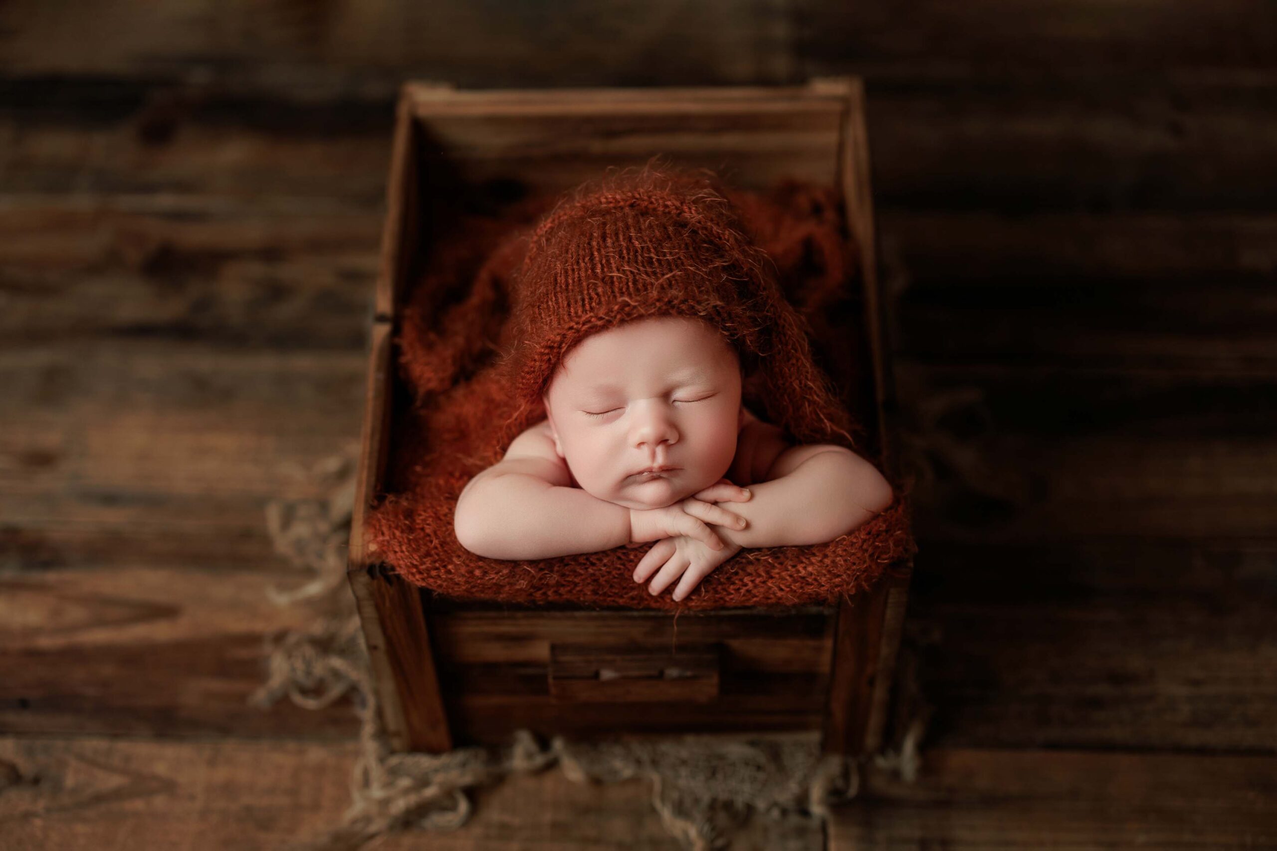A newborn baby with a cute hat sleeps peacefully with arms folded under its chin, resting on a soft brown blanket inside a rustic wooden box on a wooden floor—capturing the charm of Adelaide baby photos.