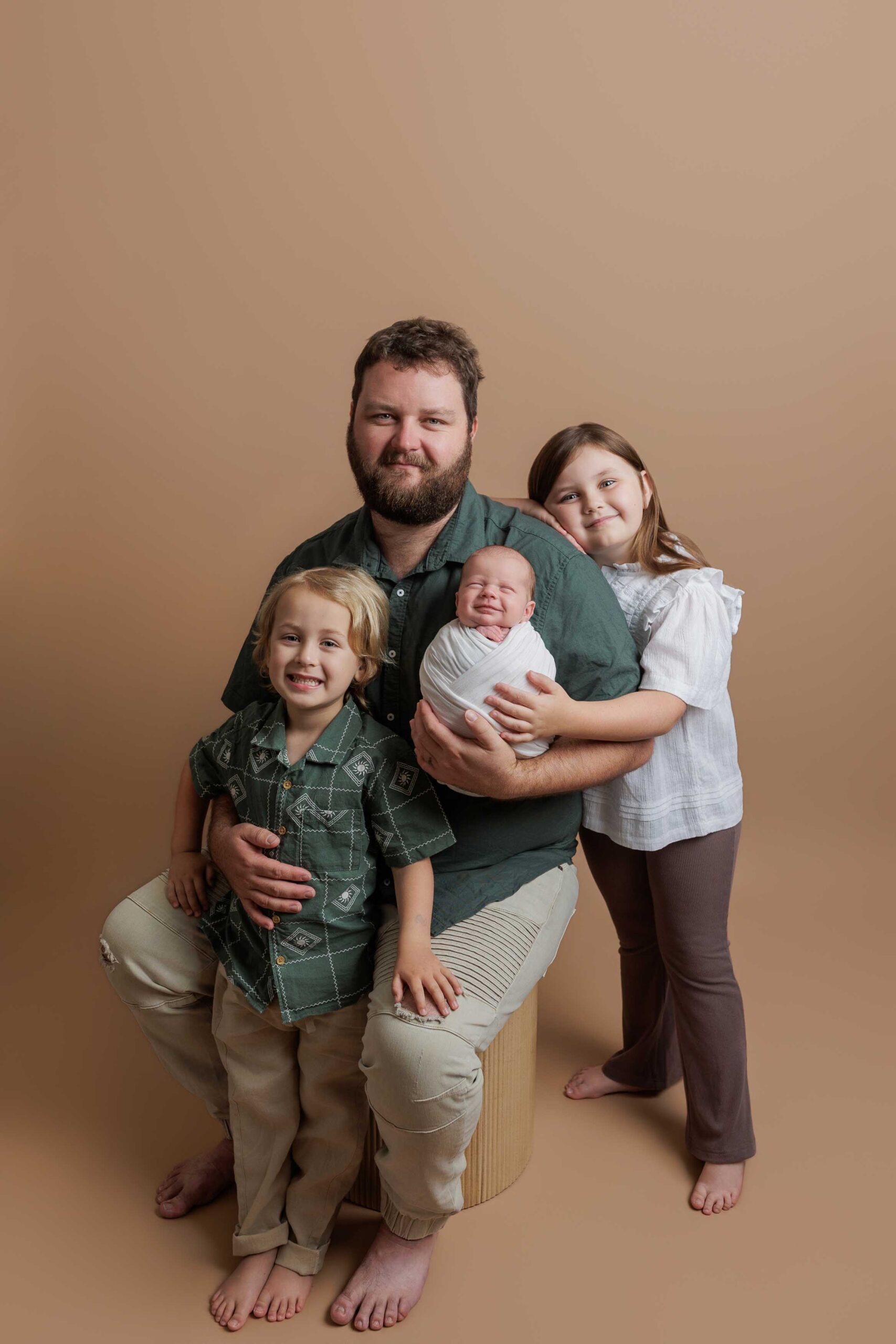 A bearded man sits on a stool holding a newborn baby, with a young boy smiling beside him and an older girl supporting the baby. All are barefoot against a plain beige background—capturing precious Adelaide baby photos with family.