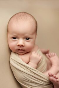 An awake Adelaide baby wrapped in a beige wrap laying on a beige background is looking at the camera.
