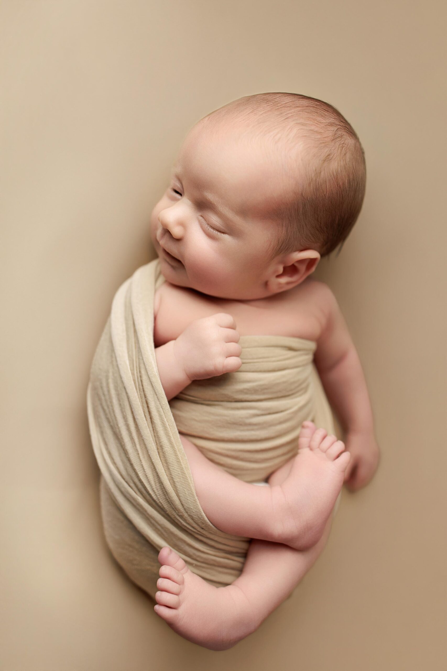 A top view of an awake Adelaide baby wrapped in a beige wrap laying on a beige background is looking at the camera.