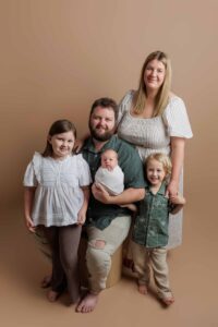 A family of five poses against a beige background in this Adelaide baby photos session. The father sits holding a swaddled baby, with a young girl to his left and a young boy to his right, whilst the mother stands behind them, smiling.