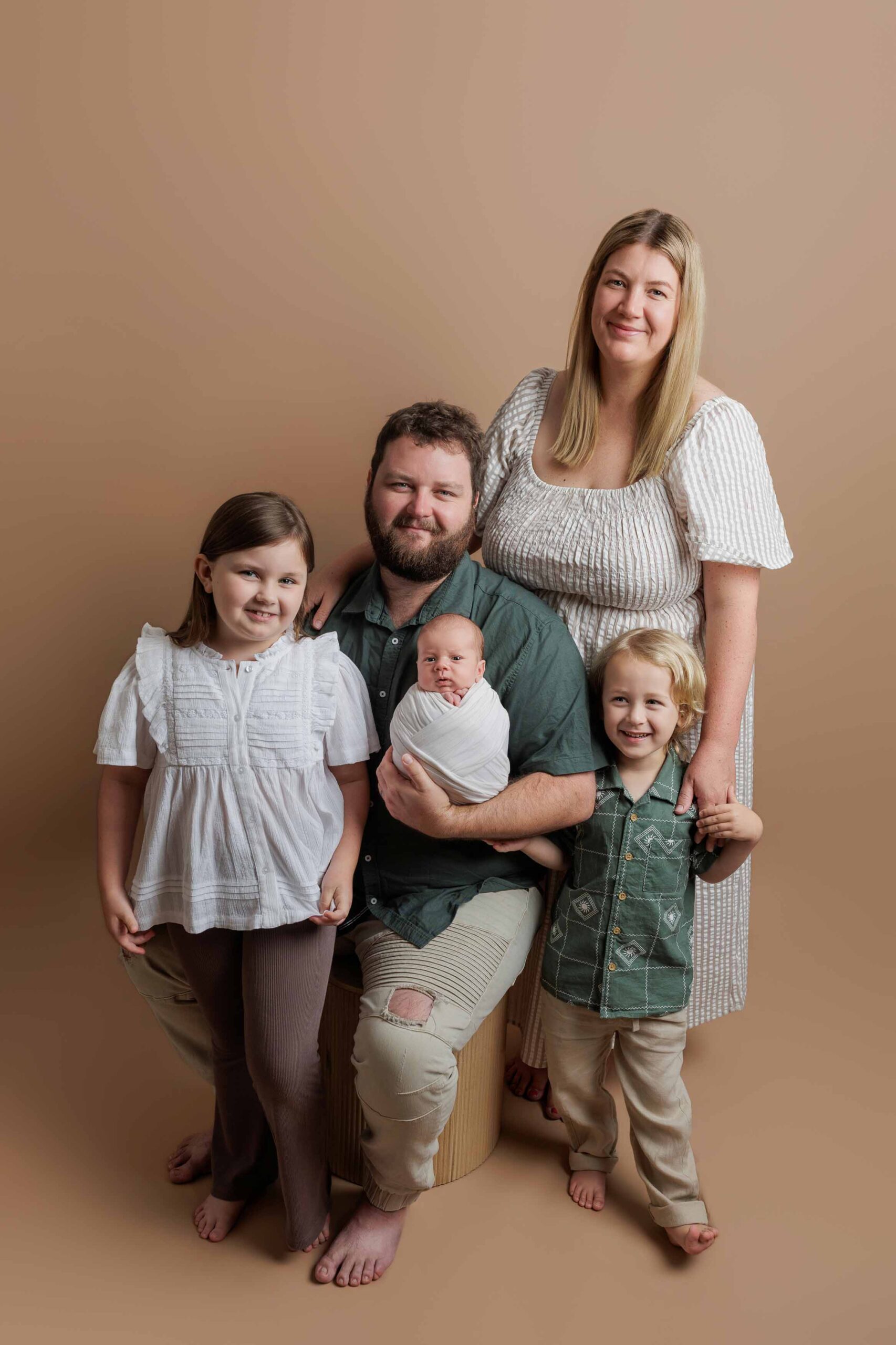 A family of five poses against a beige background in this Adelaide baby photos session. The father sits holding a swaddled baby, with a young girl to his left and a young boy to his right, whilst the mother stands behind them, smiling.