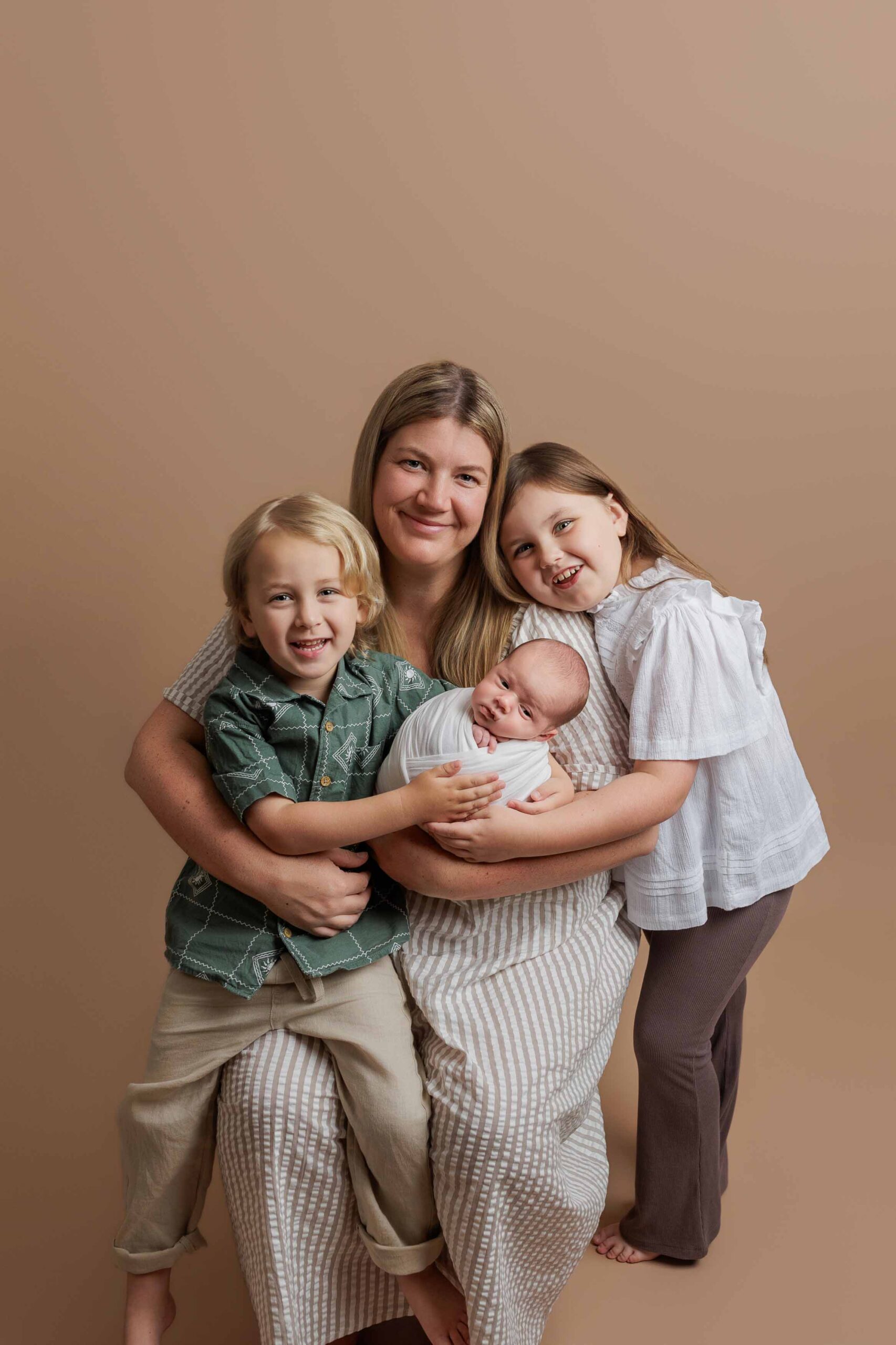 A woman smiles whilst sitting and holding a swaddled baby in her lap, with a young boy beside her and a young girl standing nearby—all captured in timeless Adelaide baby photographs against a plain beige background.