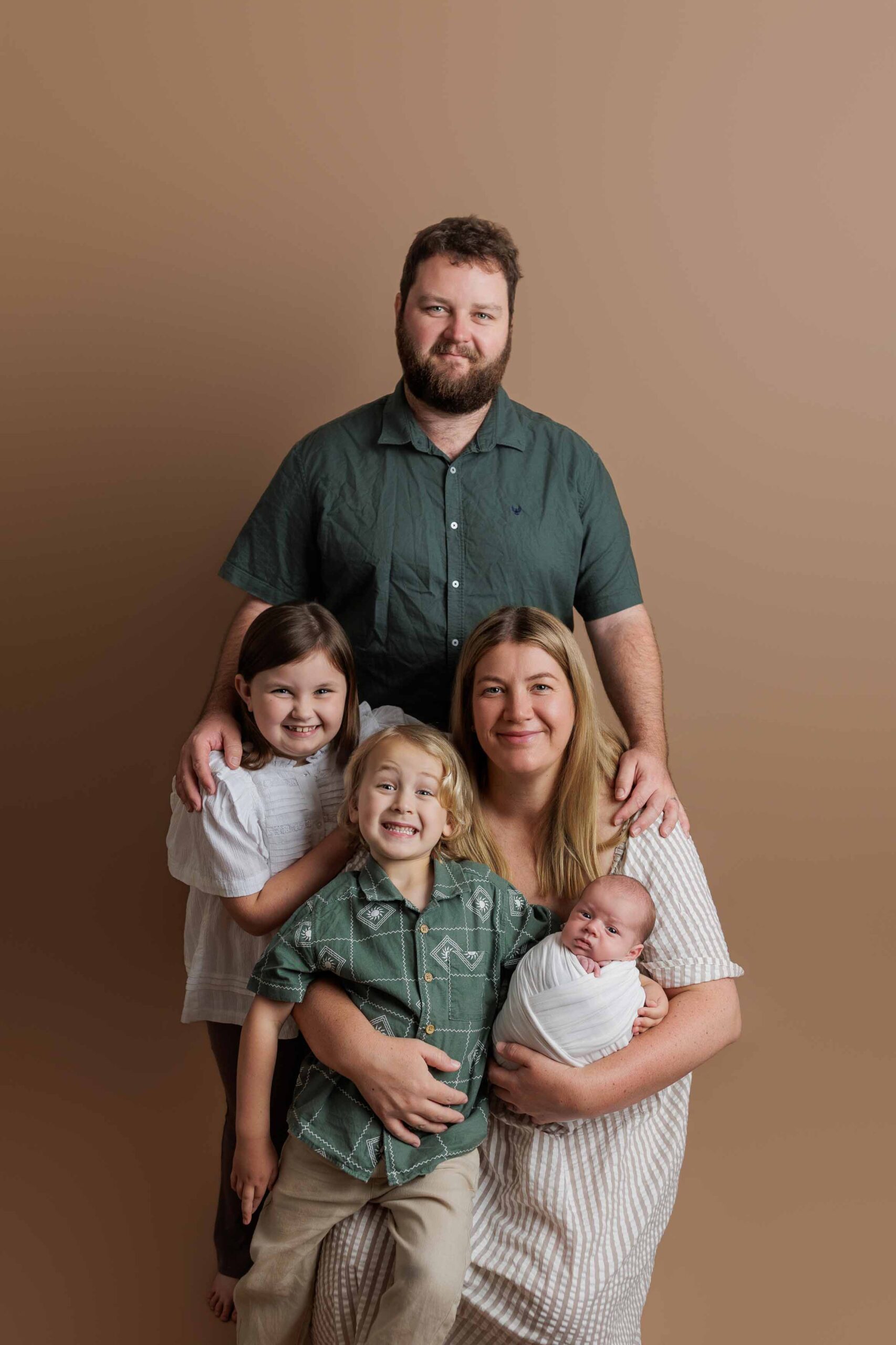 A family of five poses together in front of a plain brown background. Perfect for Adelaide baby photos, an adult man stands behind an adult woman holding a swaddled baby, with two young children smiling beside and in front of them.