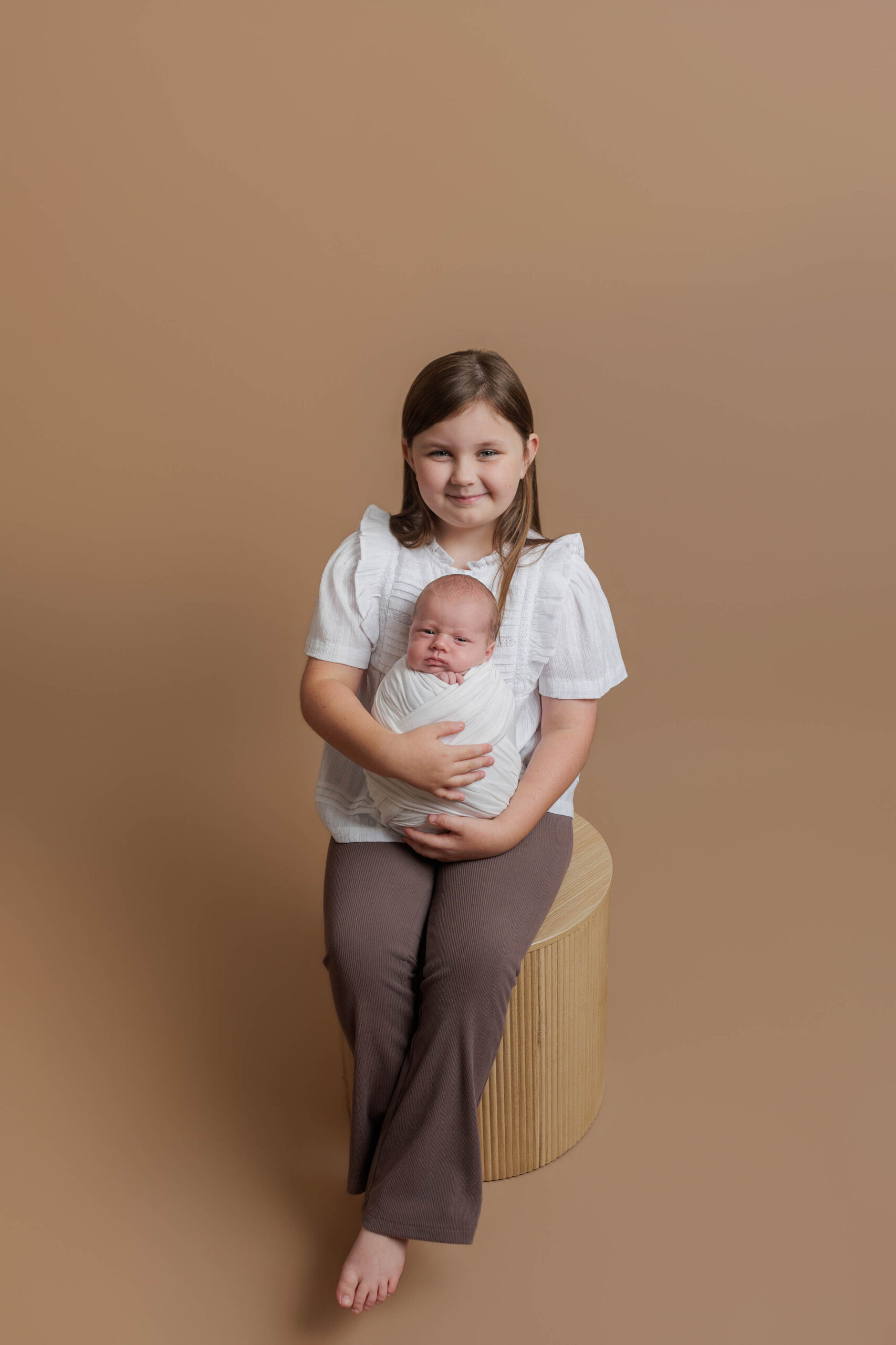 A young girl sits on a wooden stool, smiling and holding a swaddled newborn baby in her arms against a plain beige background—capturing the warmth of Adelaide baby photos.