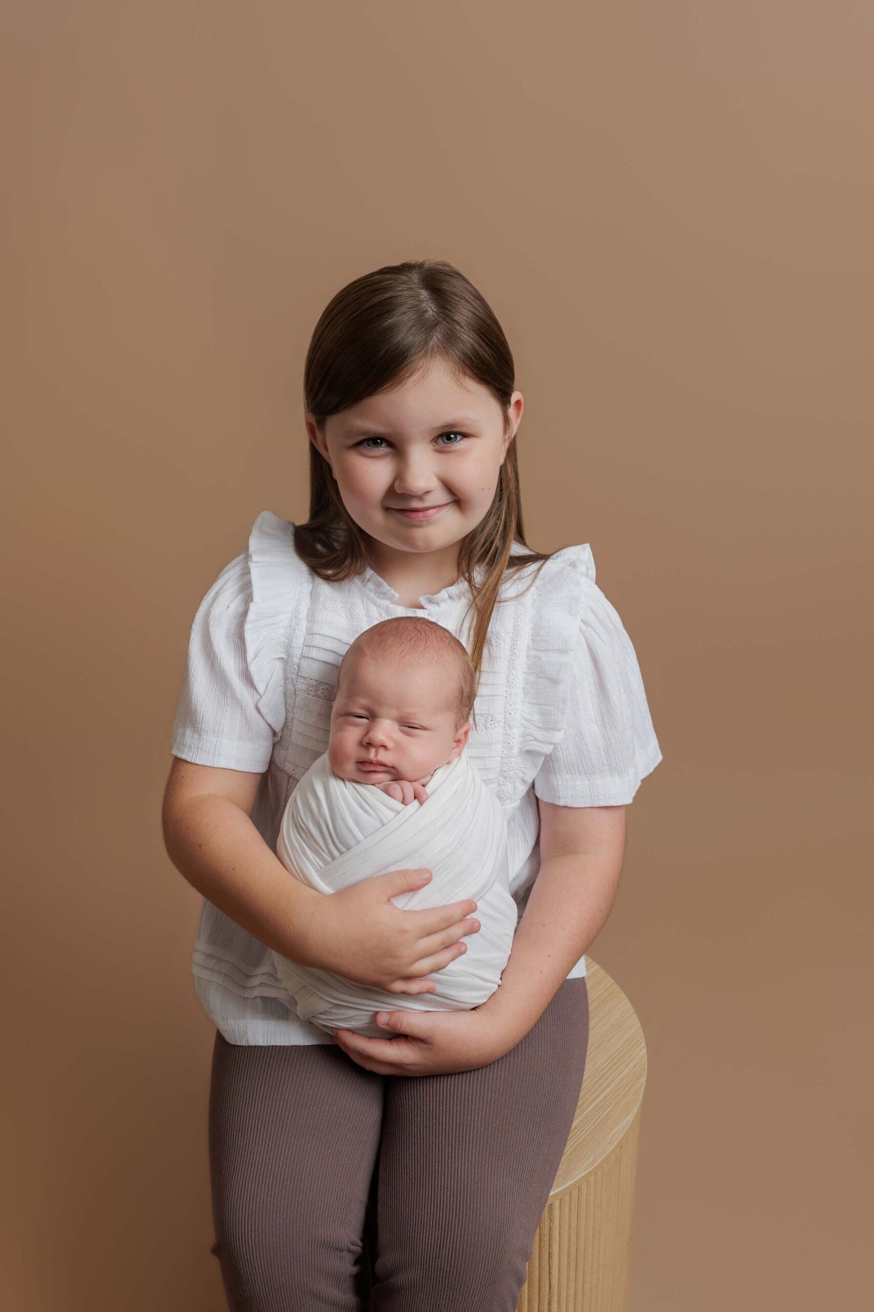 A young girl with brown hair sits on a stool, smiling and holding a newborn baby swaddled in a white blanket against a plain beige background, capturing the warmth of Adelaide baby photos.