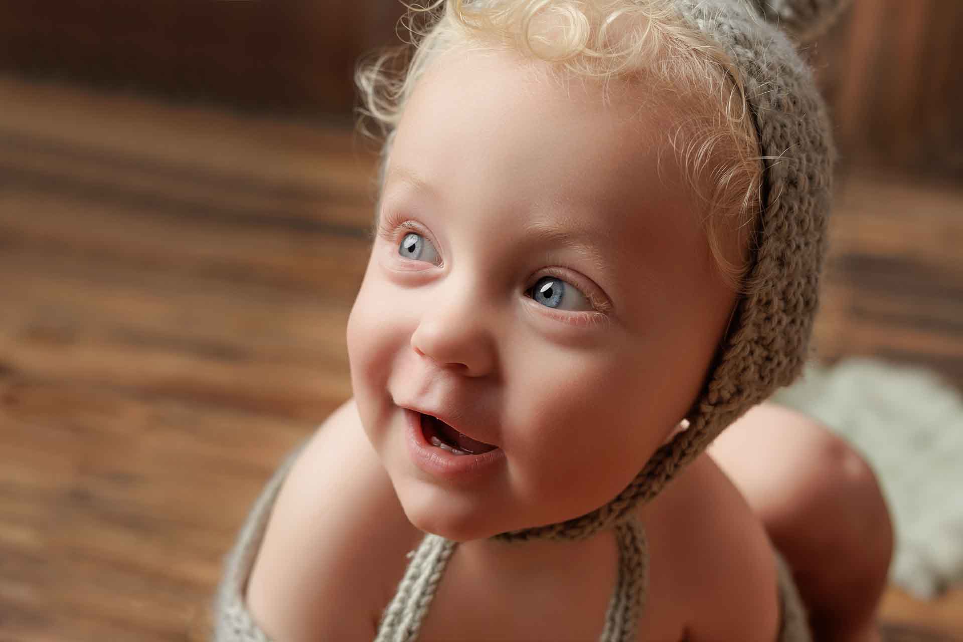 A smiling baby with curly blond hair and blue eyes wears a knitted bonnet, looking up and to the side. The background is blurred and wooden, creating a warm, cosy atmosphere—perfect for charming Adelaide baby photos.
