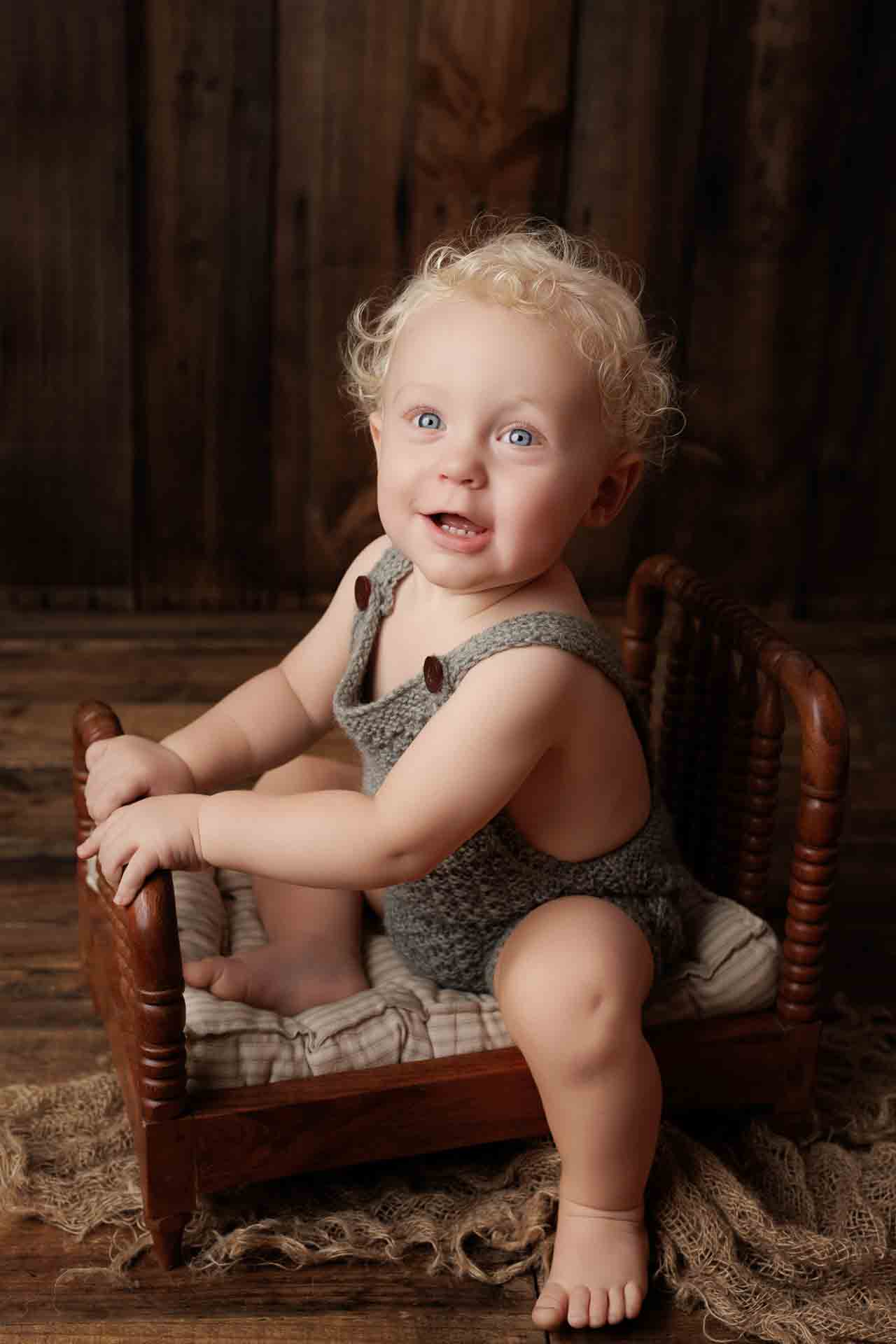 A smiling toddler with curly blonde hair sits on a small wooden bed, wearing a knitted grey romper. The rustic wooden walls and floor create a cosy setting, perfect for charming Adelaide baby photos.