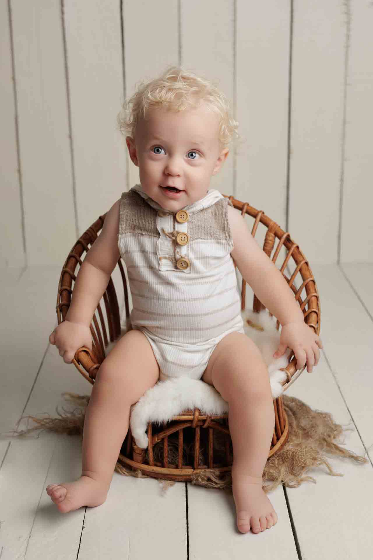 A smiling baby with curly blonde hair sits in a small wicker chair, wearing a striped sleeveless playsuit with wooden buttons, against a white wooden background—perfect for Adelaide baby photos.