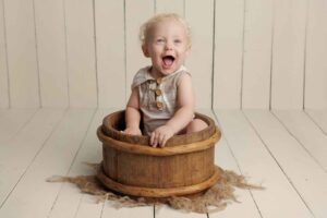 A smiling baby with curly blonde hair sits inside a rustic wooden tub on a hessian cloth, against a white wooden floor and wall background—perfect for capturing Adelaide baby photos.