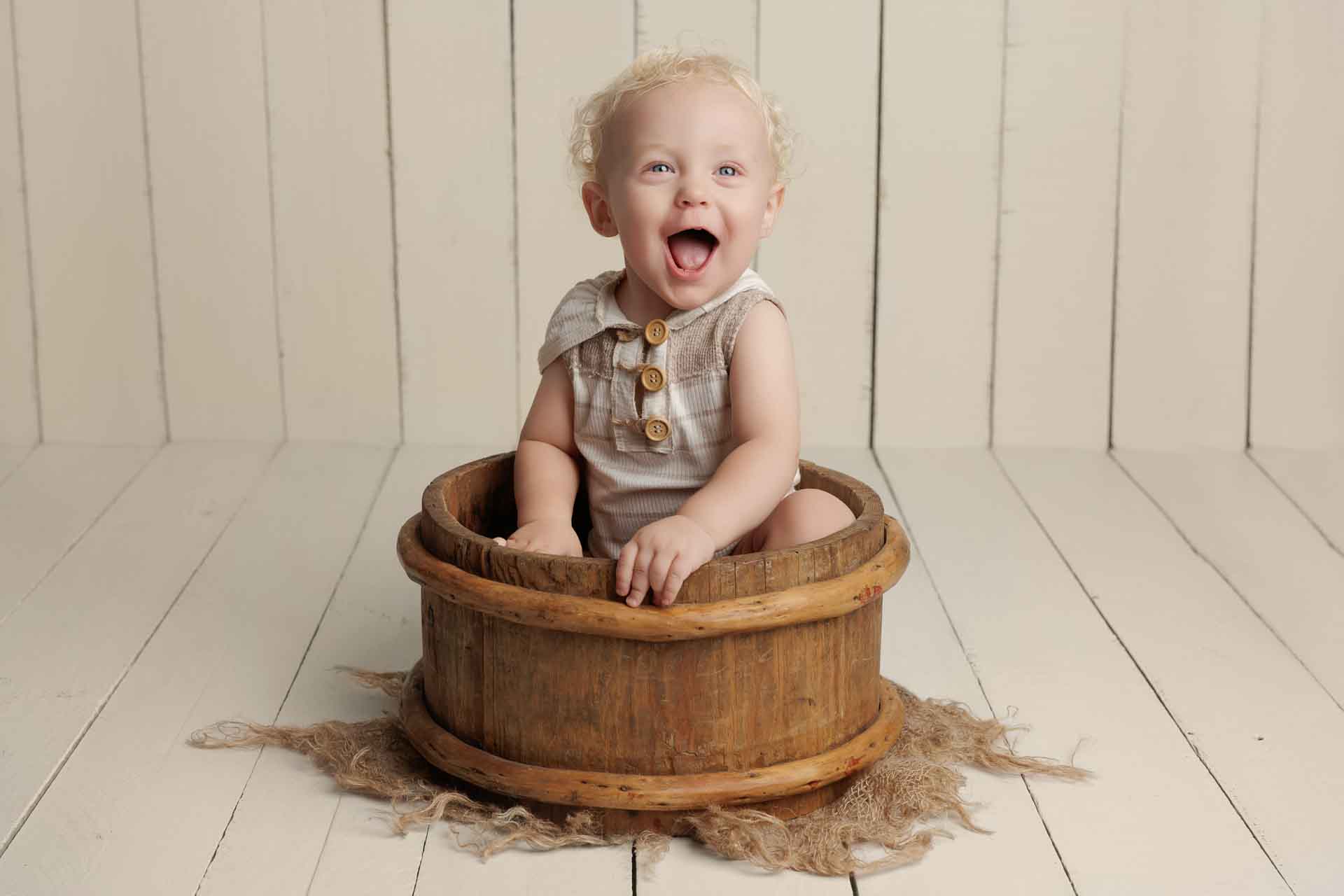 A smiling baby with curly blonde hair sits inside a rustic wooden tub on a hessian cloth, against a white wooden floor and wall background—perfect for capturing Adelaide baby photos.