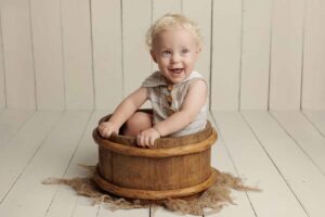 A smiling baby with curly blonde hair sits inside a rustic wooden tub on a light wooden floor, wearing a light-coloured outfit with buttons—capturing the pure joy of Adelaide baby photos.