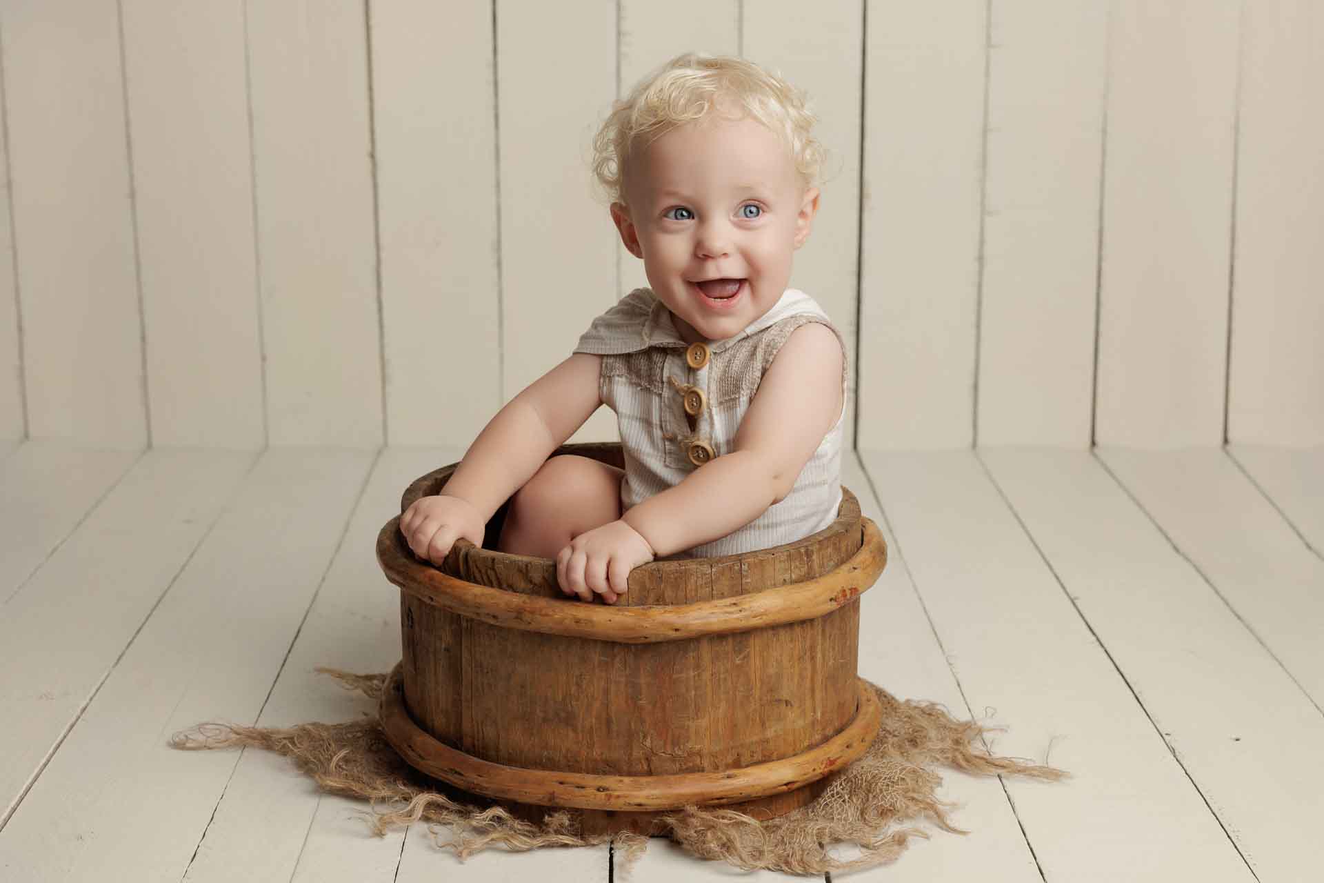 A smiling baby with curly blonde hair sits inside a rustic wooden tub on a light wooden floor, wearing a light-coloured outfit with buttons—capturing the pure joy of Adelaide baby photos.