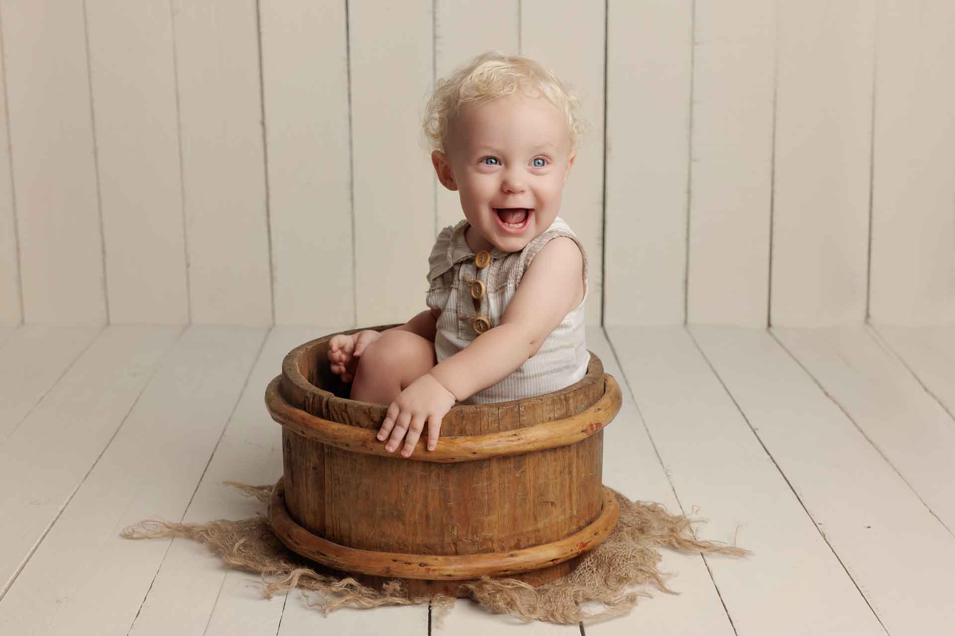 A smiling baby with curly blonde hair sits inside a rustic wooden tub on a wooden floor, wearing a light-coloured outfit. The simple background with vertical wooden panels creates a charming scene perfect for Adelaide baby photos.