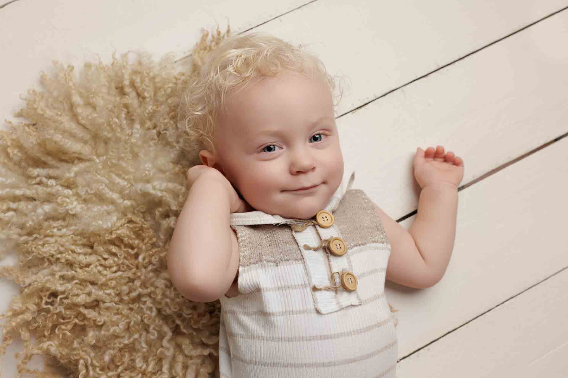 A smiling toddler with curly blonde hair lies on a cream-coloured textured rug and white wooden floor, wearing a sleeveless shirt with buttons—perfect for Adelaide baby photos.