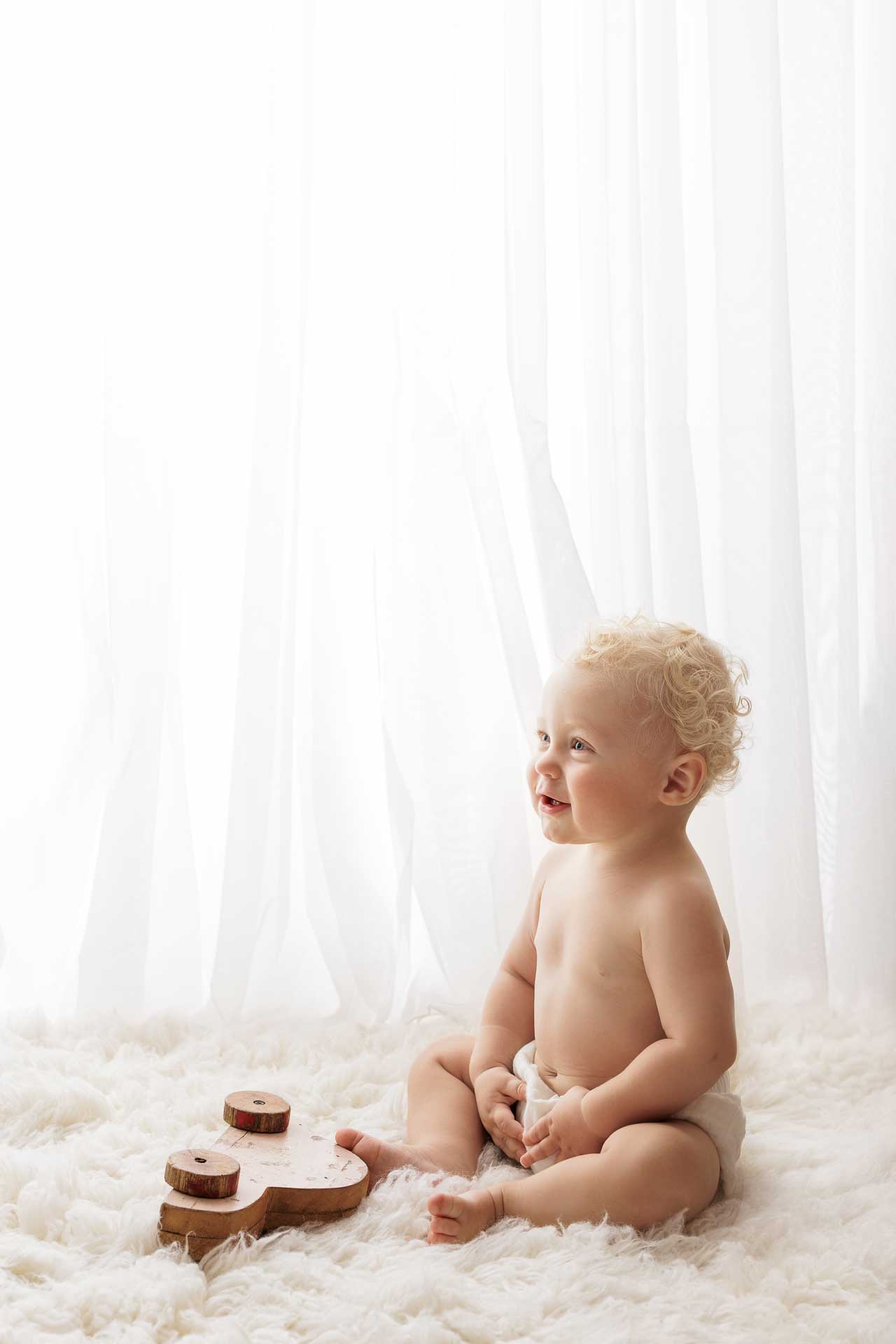 A smiling baby with curly blonde hair sits on a soft white rug in front of sheer curtains, wearing only a nappy. A wooden number 2 is placed on the rug beside the baby, capturing a sweet moment perfect for Adelaide baby photos.