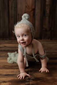 A smiling baby with curly blonde hair, wearing a knitted bonnet and outfit, crawls on a wooden floor with a soft green blanket in the background—perfect for adorable Adelaide baby photos.