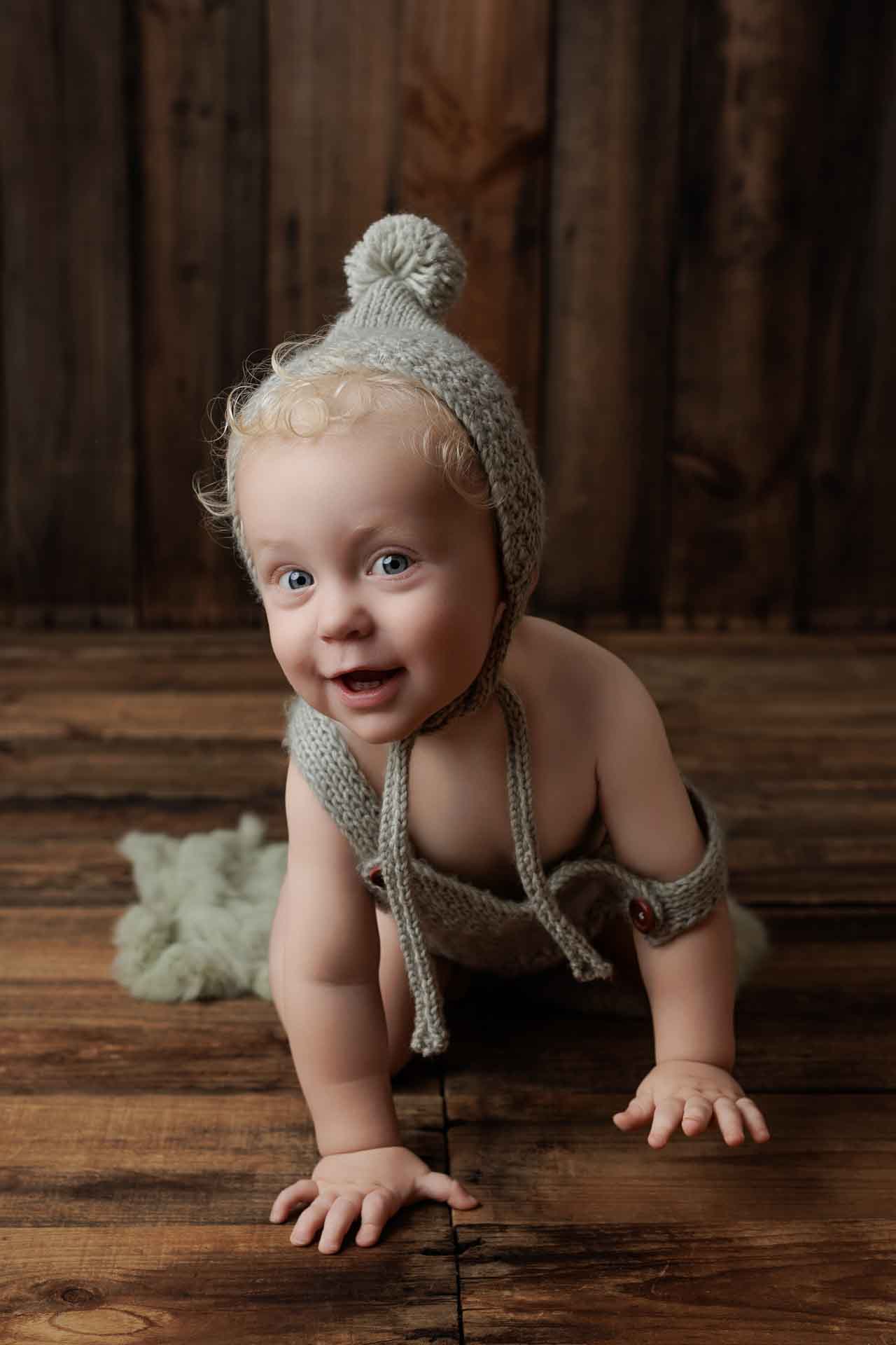 A smiling baby with curly blonde hair, wearing a knitted bonnet and outfit, crawls on a wooden floor with a soft green blanket in the background—perfect for adorable Adelaide baby photos.