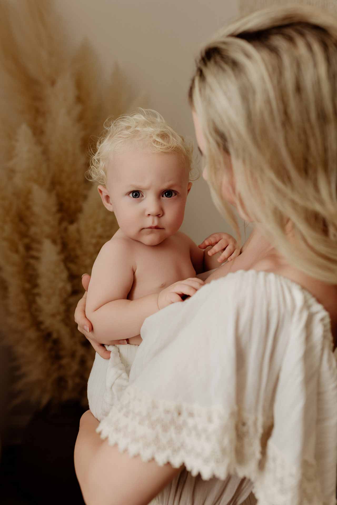 A woman with blonde hair in a white dress holds a blonde, blue-eyed baby who looks directly at the camera, creating a timeless moment. The neutral background with tall pampas grass décor makes this perfect for Adelaide baby photos.