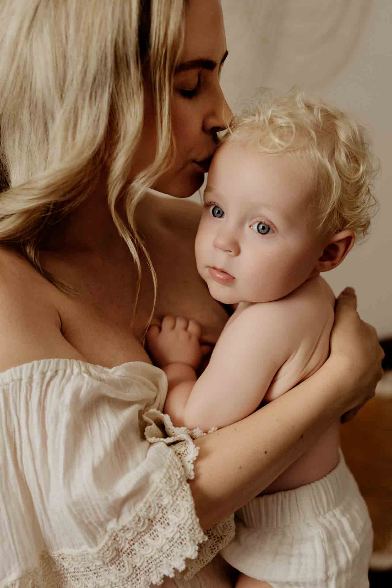 A woman with long blonde hair kisses the forehead of a young, curly-haired baby in her arms. Dressed in light-coloured clothing, they share a tender, loving moment—perfectly capturing the warmth of Adelaide baby photos.