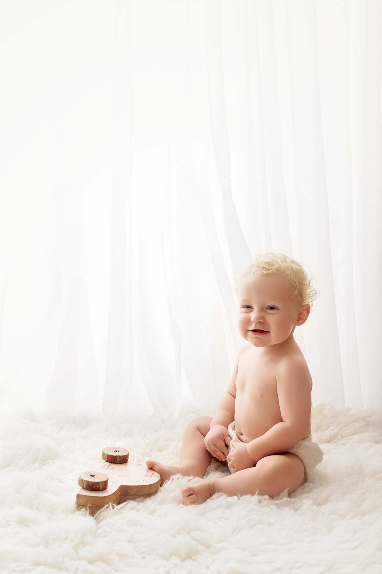 A smiling baby with curly blonde hair sits on a fluffy white rug, wearing only a nappy. A wooden number 1 is placed next to the baby, capturing a sweet moment for Adelaide baby photos with sheer white curtains in the background.