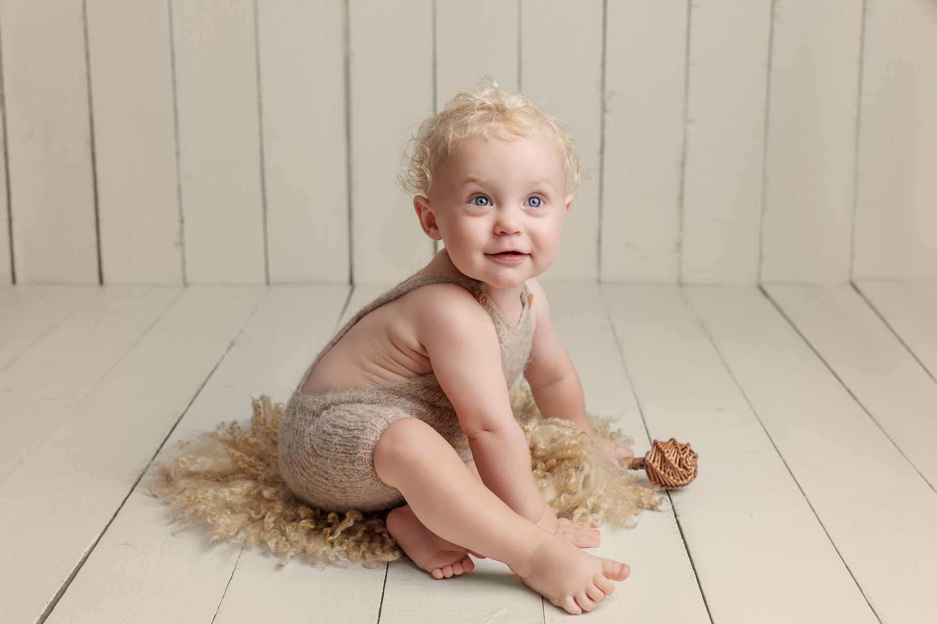 A smiling baby with curly blonde hair sits on a fluffy beige rug, wearing a knitted beige romper, on a light wooden floor with white wooden panelled walls in the background. Perfect for Adelaide baby photos, a small pine cone is nearby.
