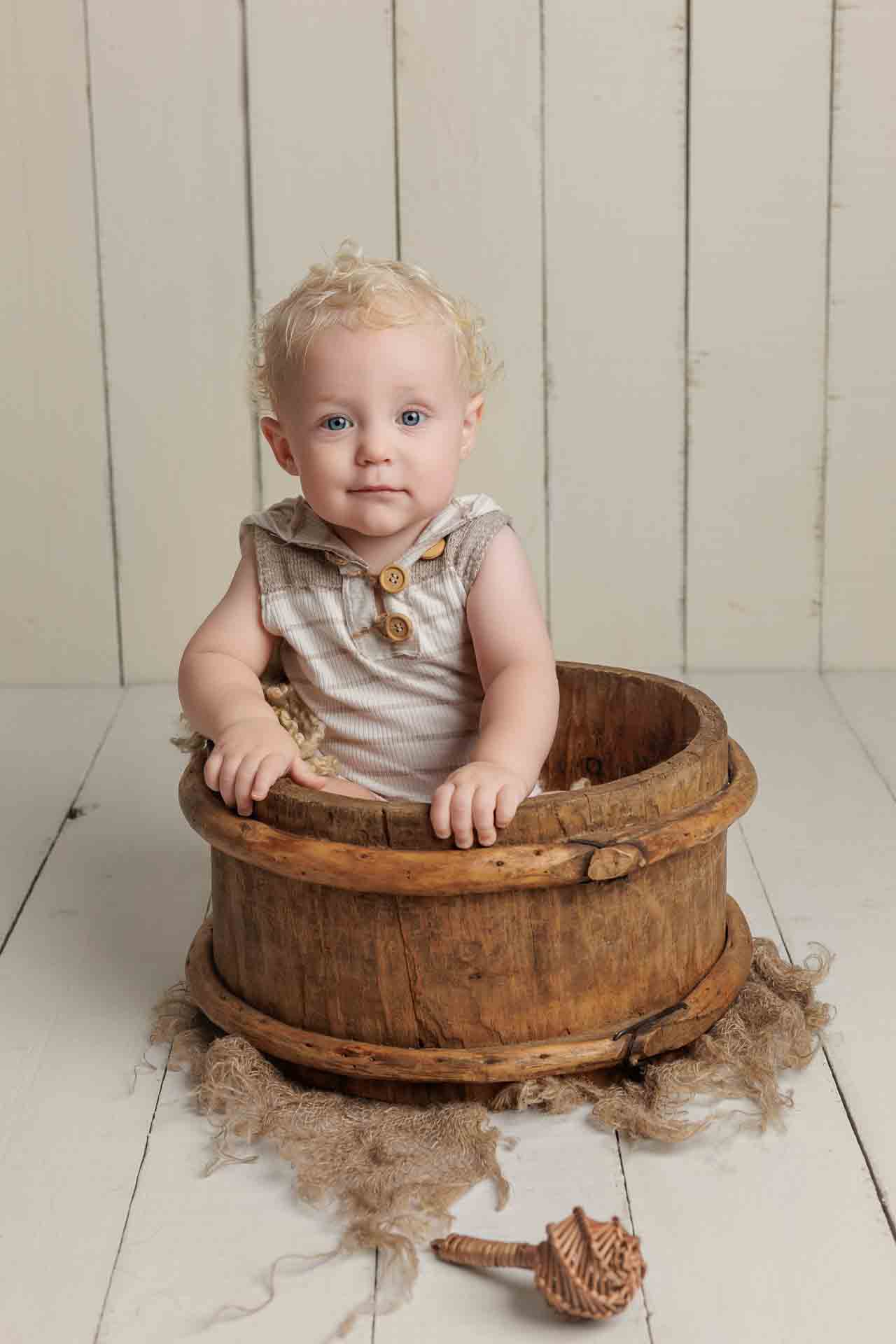 A blonde baby with blue eyes sits inside a rustic wooden barrel on a wooden floor, wearing a light sleeveless outfit. Perfect for Adelaide baby photos, with a woven toy and hessian cloth in front and white wooden panels as the backdrop.