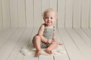 Smiling toddler with curly blonde hair sits on a fluffy white rug in an Adelaide baby photography session, wearing a green and white checked romper against a light wooden panelled background.