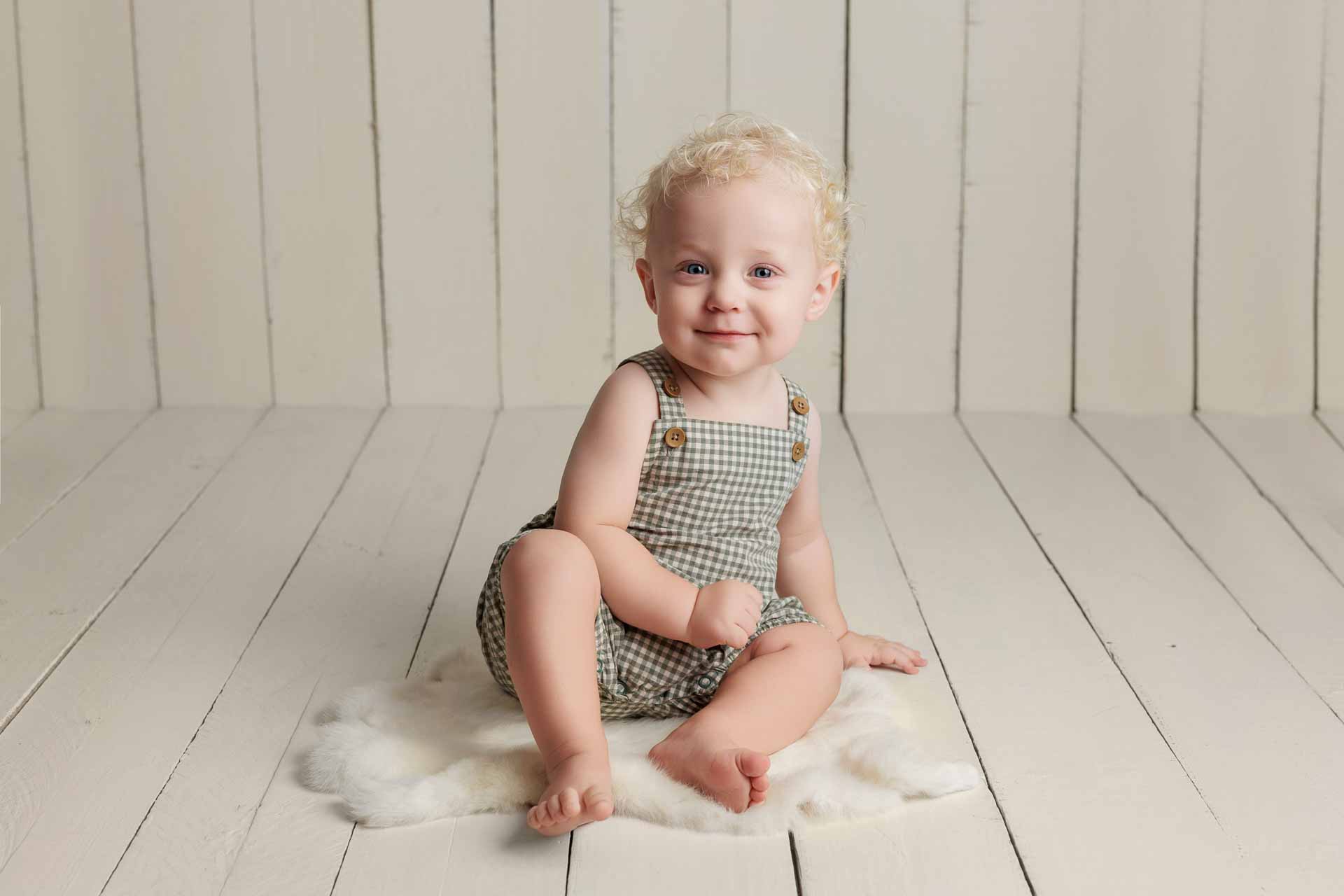 Smiling toddler with curly blonde hair sits on a fluffy white rug in an Adelaide baby photography session, wearing a green and white checked romper against a light wooden panelled background.