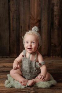 A smiling baby with curly blonde hair sits on a soft green rug, wearing a knitted grey romper and matching pom-pom hat, perfect for Adelaide baby photos against a rustic wooden background.