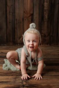 A smiling baby wearing a knitted grey outfit and pom-pom hat crouches on a fuzzy green rug against a rustic wooden background—an adorable moment captured in these Adelaide baby photos.