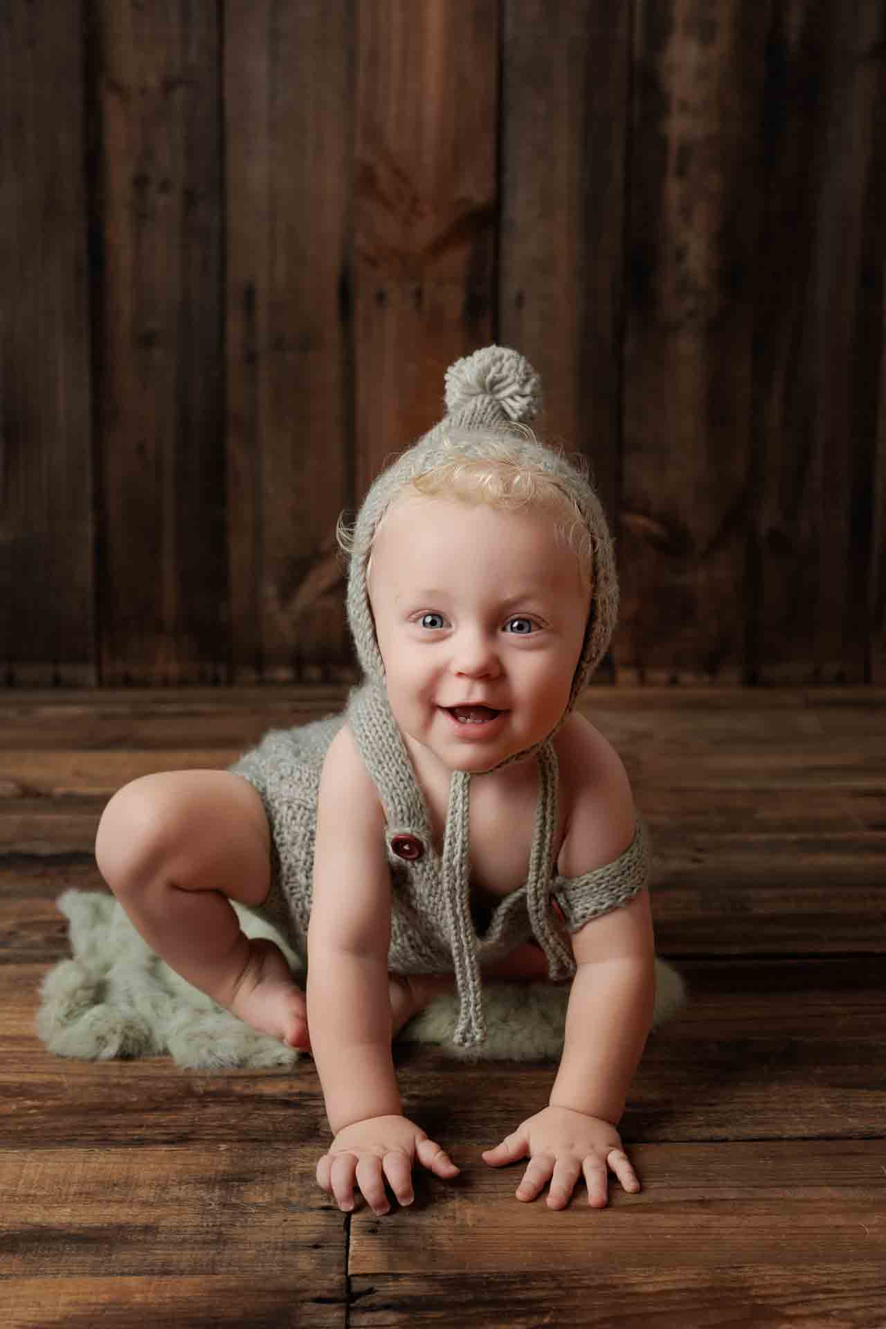 A smiling baby wearing a knitted grey outfit and pom-pom hat crouches on a fuzzy green rug against a rustic wooden background—an adorable moment captured in these Adelaide baby photos.