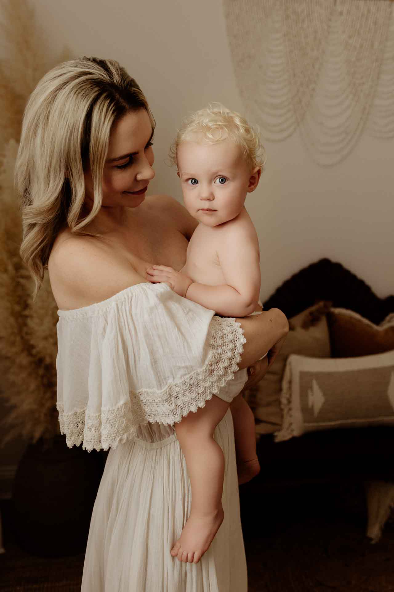 A woman in an off-the-shoulder white dress holds a blonde, blue-eyed toddler. Indoors with neutral-toned decor, she smiles gently at the child, who looks at the camera—capturing a precious Adelaide baby photos moment.